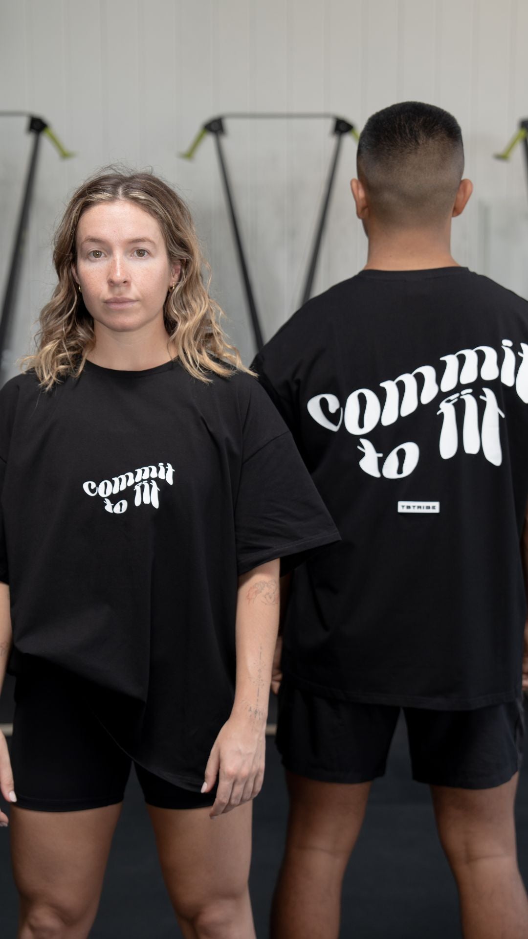 Male and Female model wearing a black oversized t shirt with white writing on the front and back that reads Commit to Fit